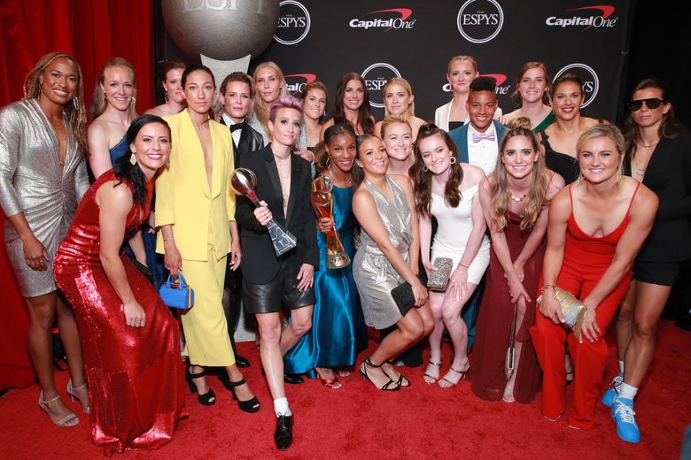 The USWNT stands on a red carpet, smiling, while Megan Rapinoe and Crystal Dunn hold trophies.