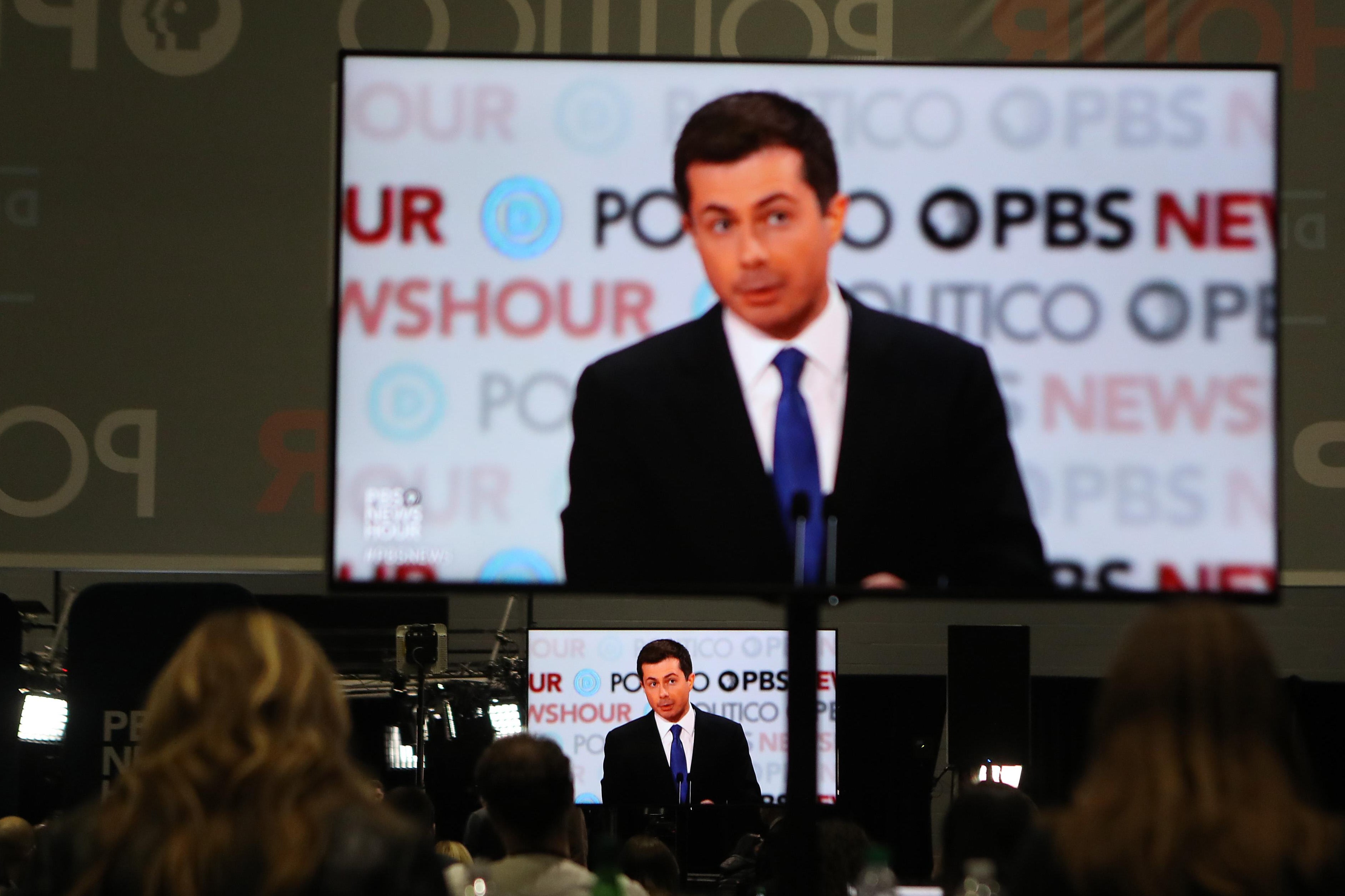Journalists, seen from behind, sit watching a large screen above a small screen, both showing the same image of South Bend, Indiana Mayor Pete Buttigieg against the debate backdrop covered with the logos of the sponsoring news organizations. (Photo by Mario Tama/Getty Images)