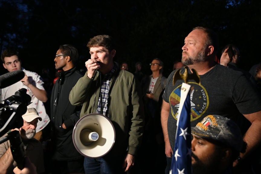 Fuentes, wearing a green jacket and standing amid a crowd, speaks into a handheld device connected to a bullhorn. Jones is standing next to him wearing a "Space Force" T-shirt.