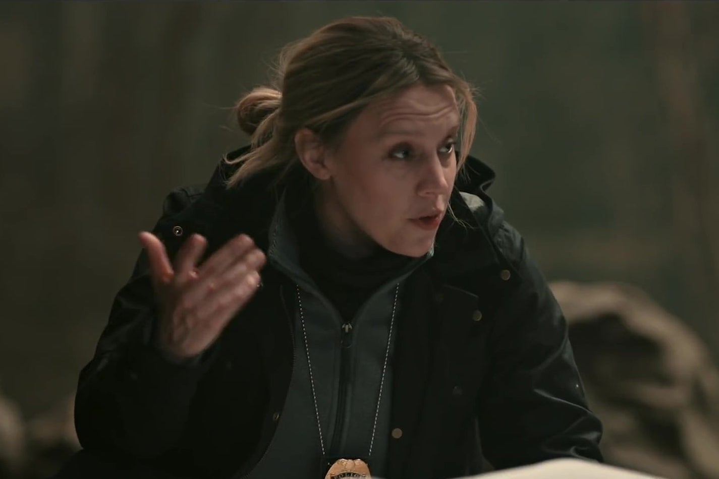 Kate McKinnon, dressed like a grizzled Pennsylvania detective, kneeling in the woods over a body covered in plastic. 