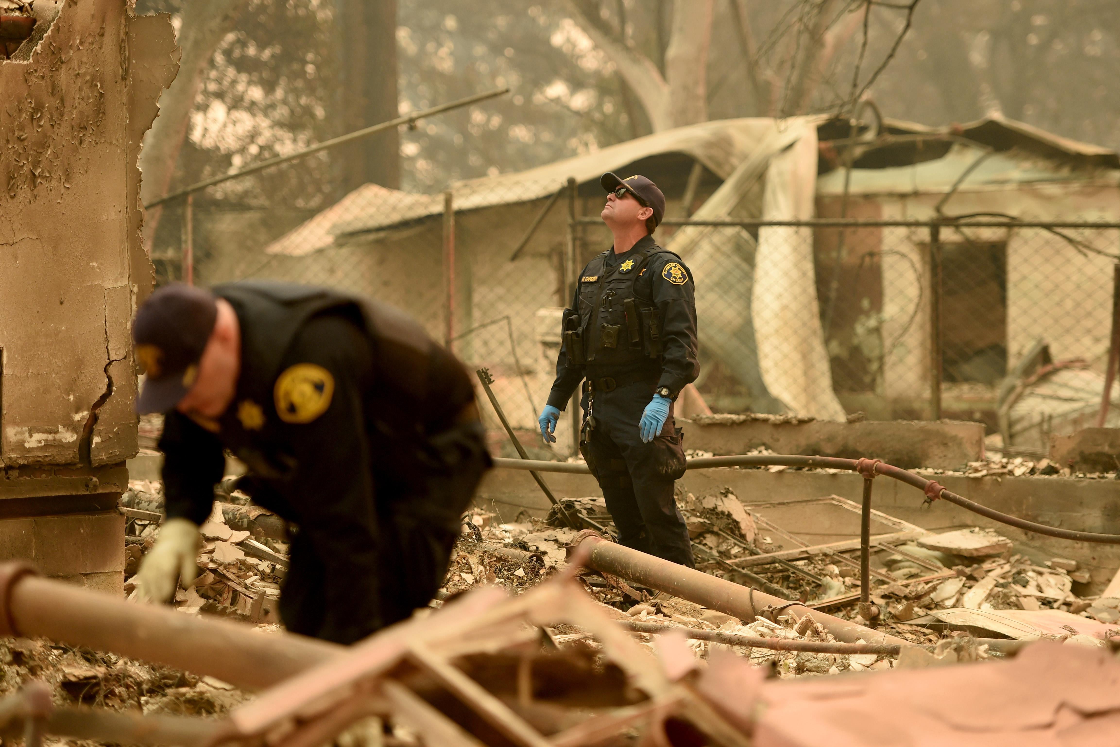 In the burned wreckage of a home, two officers wearing gloves look through the rubble. A layer of ash coats everything.