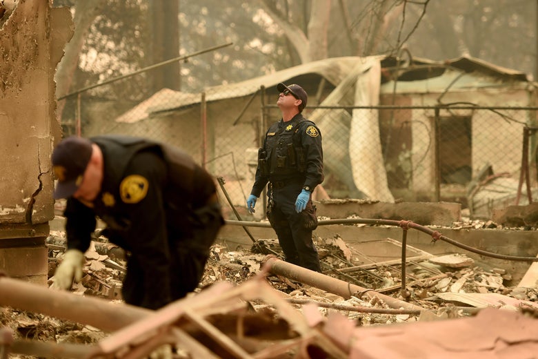 In the burned wreckage of a home, two officers wearing gloves look through the rubble. A layer of ash coats everything.