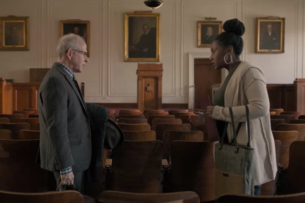 Elliot and Yaz stand in an old-looking college classroom with paintings in the front and many rows of wooden chairs.