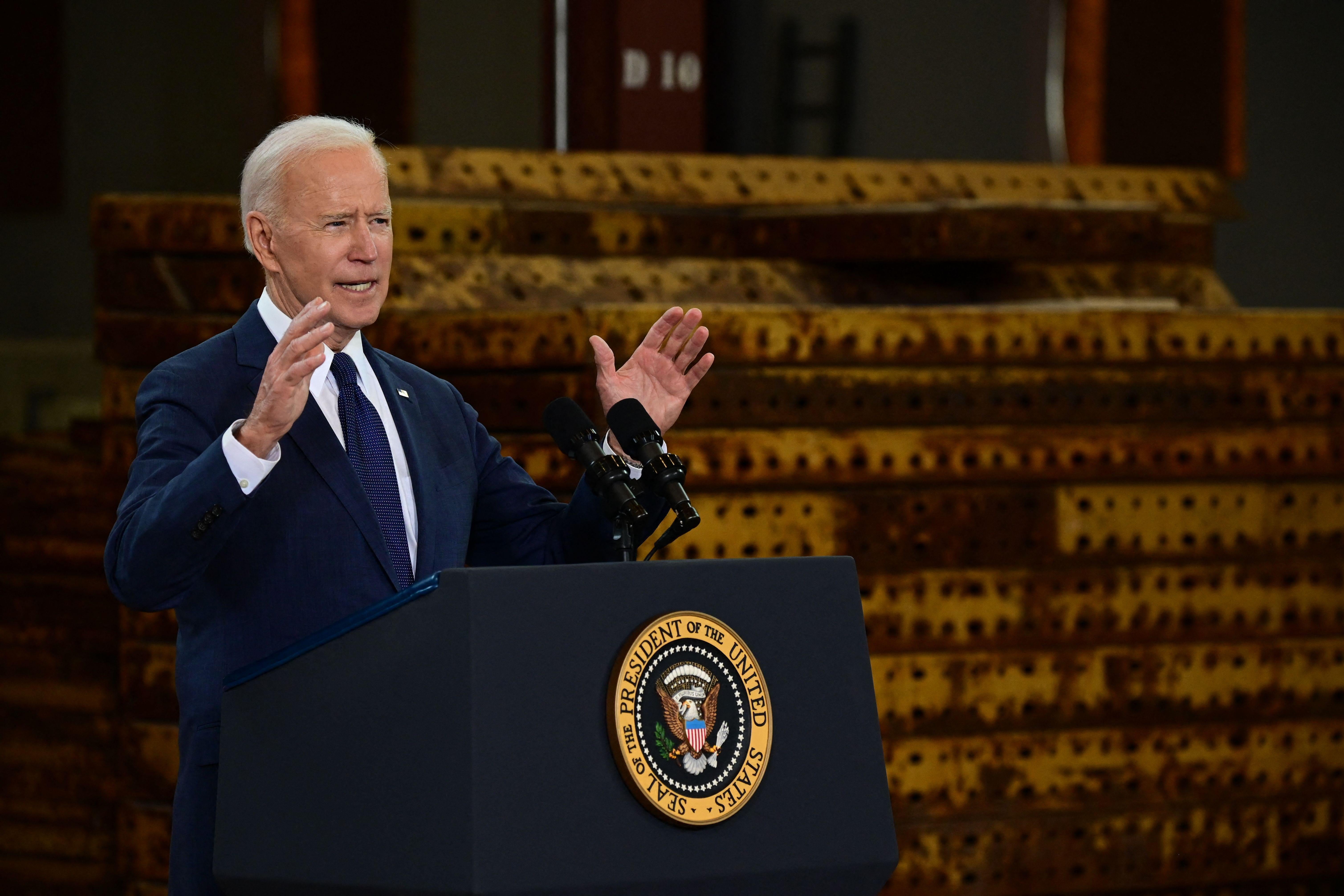 Joe Biden gestures with both hands while speaking at a podium with a stack of rusted metal behind him at a facility in Pittsburgh