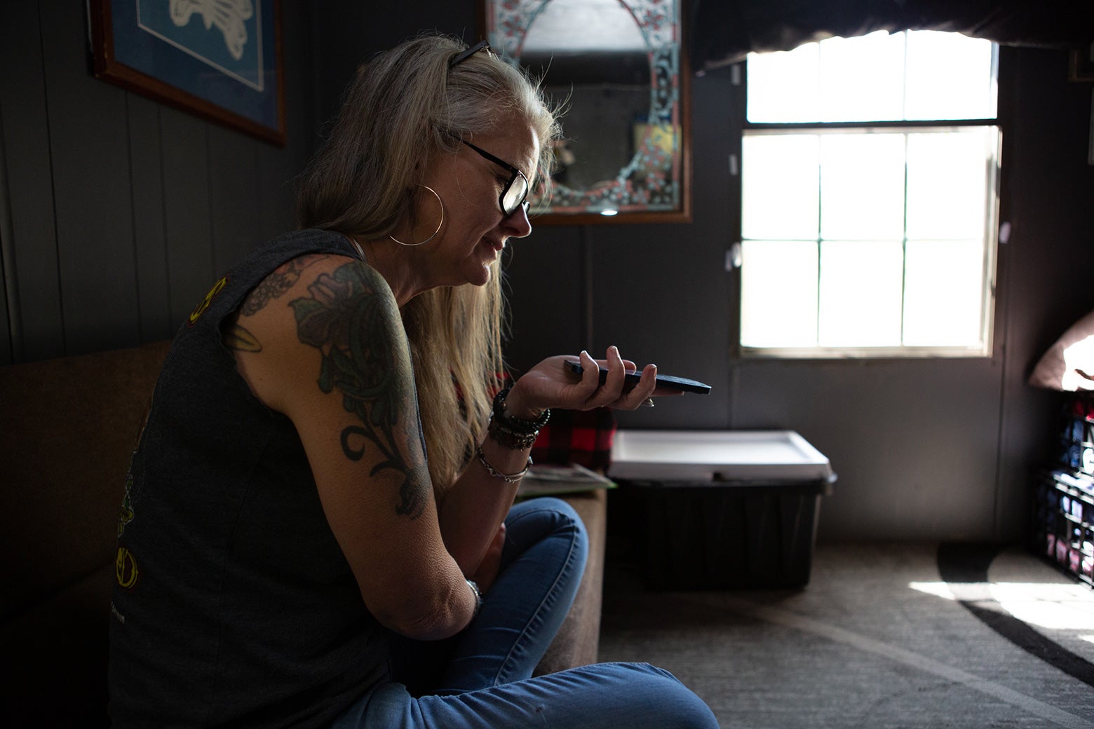 A white woman with long gray hair wearing glasses sits on a couch in a living room and talks into a phone on speakerphone.