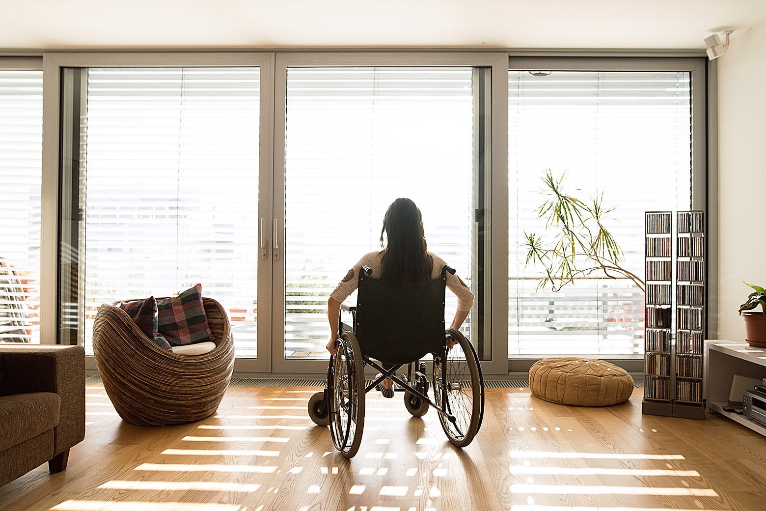 A woman in a wheelchair looking at sliding glass doors with the blinds partly open in her home.