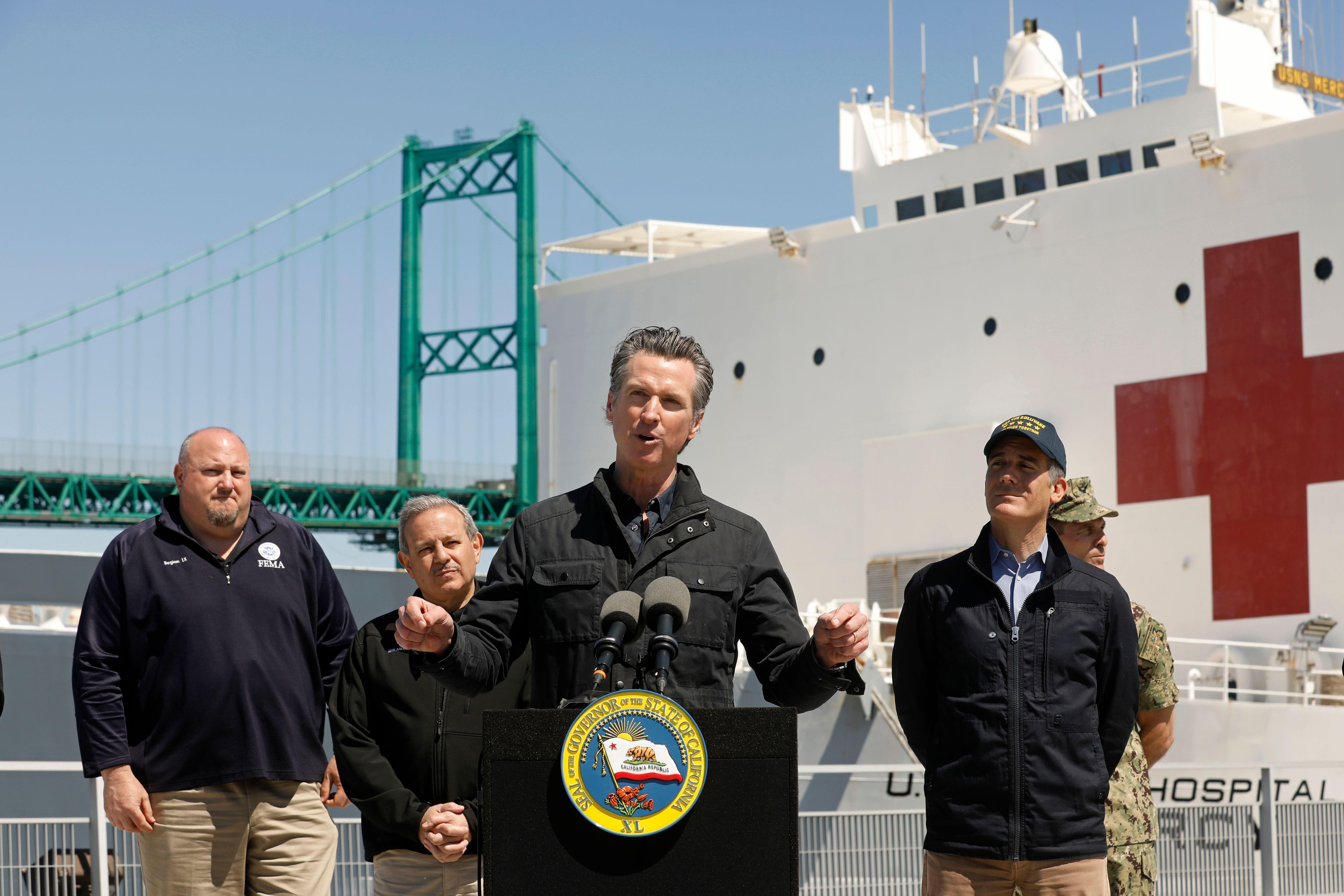 Newsom speaks at a podium with federal and local officials standing near him and a big military ship with a red cross on it behind him