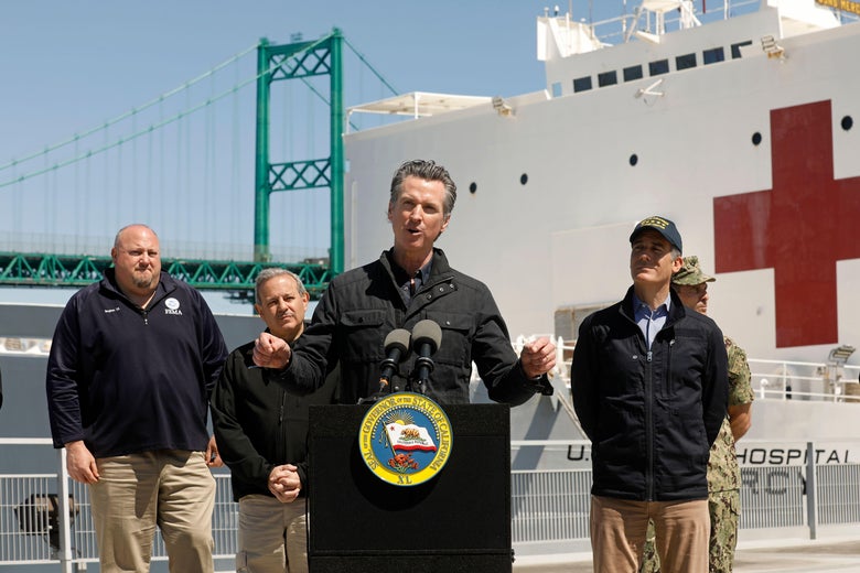 Newsom speaks at a podium with federal and local officials standing near him and a big military ship with a red cross on it behind him