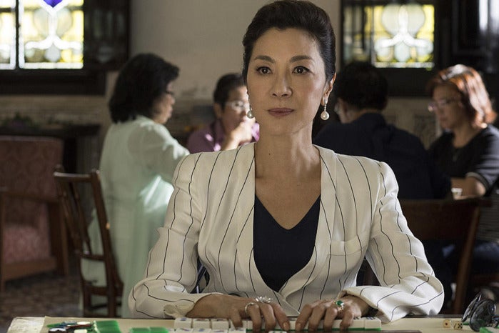 Michelle Yeoh, wearing a white pinstripe blazer, sits at a table, handling mahjong tiles.