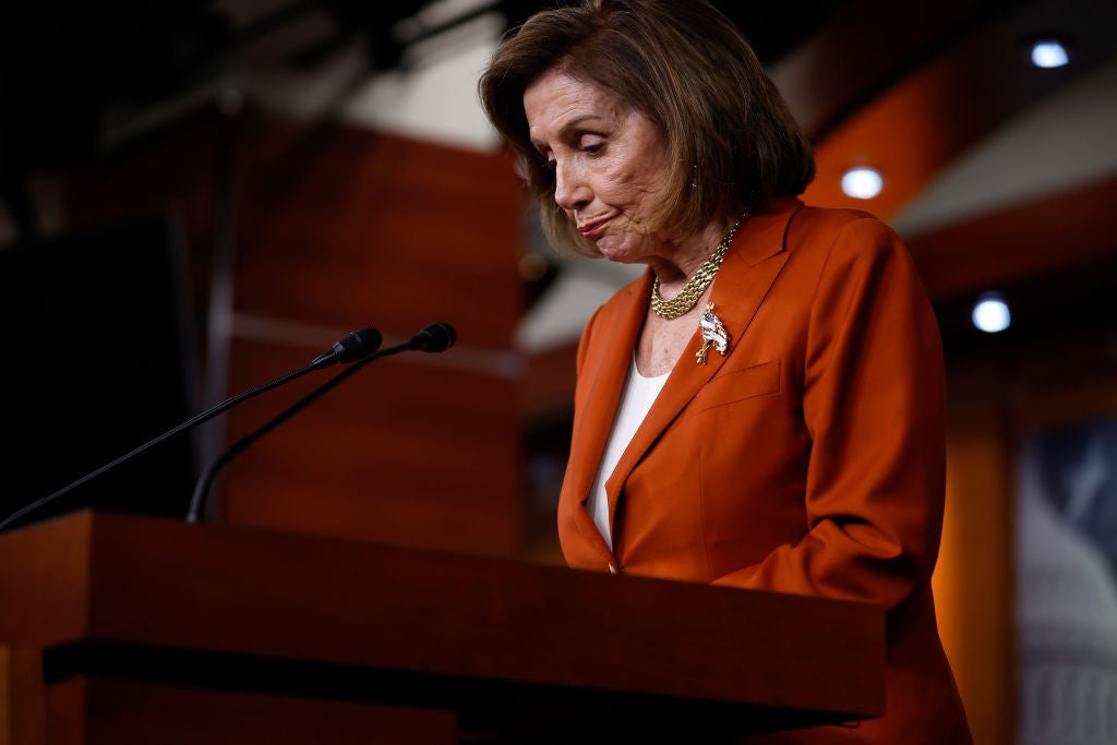 Pelosi, wearing an orange blazer, glances downward at her lectern with a defeated expression.