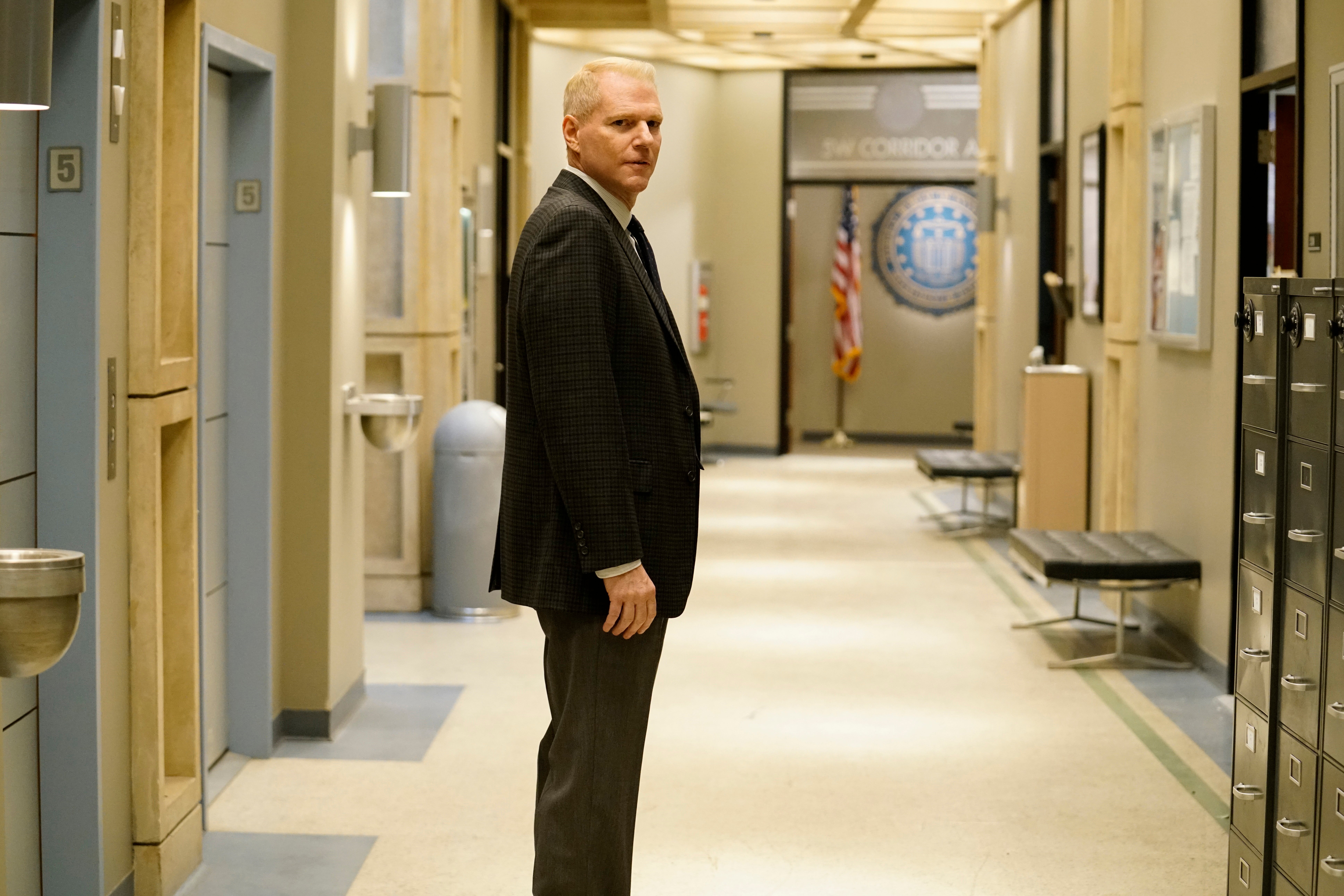 Noah Emmerich as Stan Beeman on The Americans. Emmerich is a white man in fifties with thinning blonde hair and a medium build. He wears brown trousers and a black jacket. The shot shows him from the calves up, in profile. He stands in a hallway.