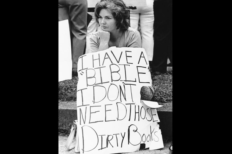 A woman sits on the curb holding a sign that says, "I have a 'Bible' I don't need those dirty books."