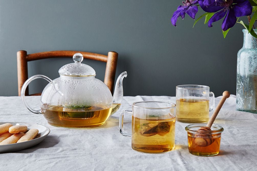 Clear glass teapot and teacups filled with golden-hued tea next to a glass honey jar with a dipper in it, a plate of tea cookies, and a flower vase on a table