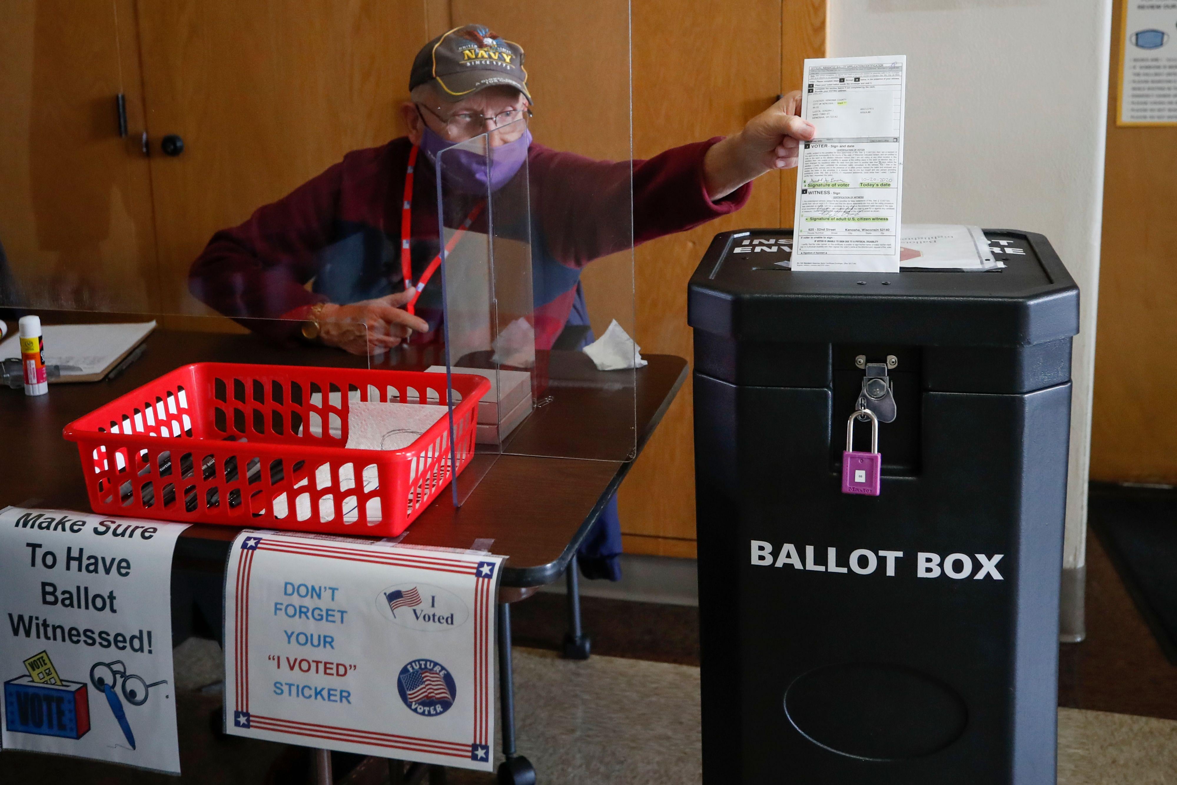 An election worker wearing a mask reaches across the table he's sitting at to drop a ballot in the slot of a ballot box