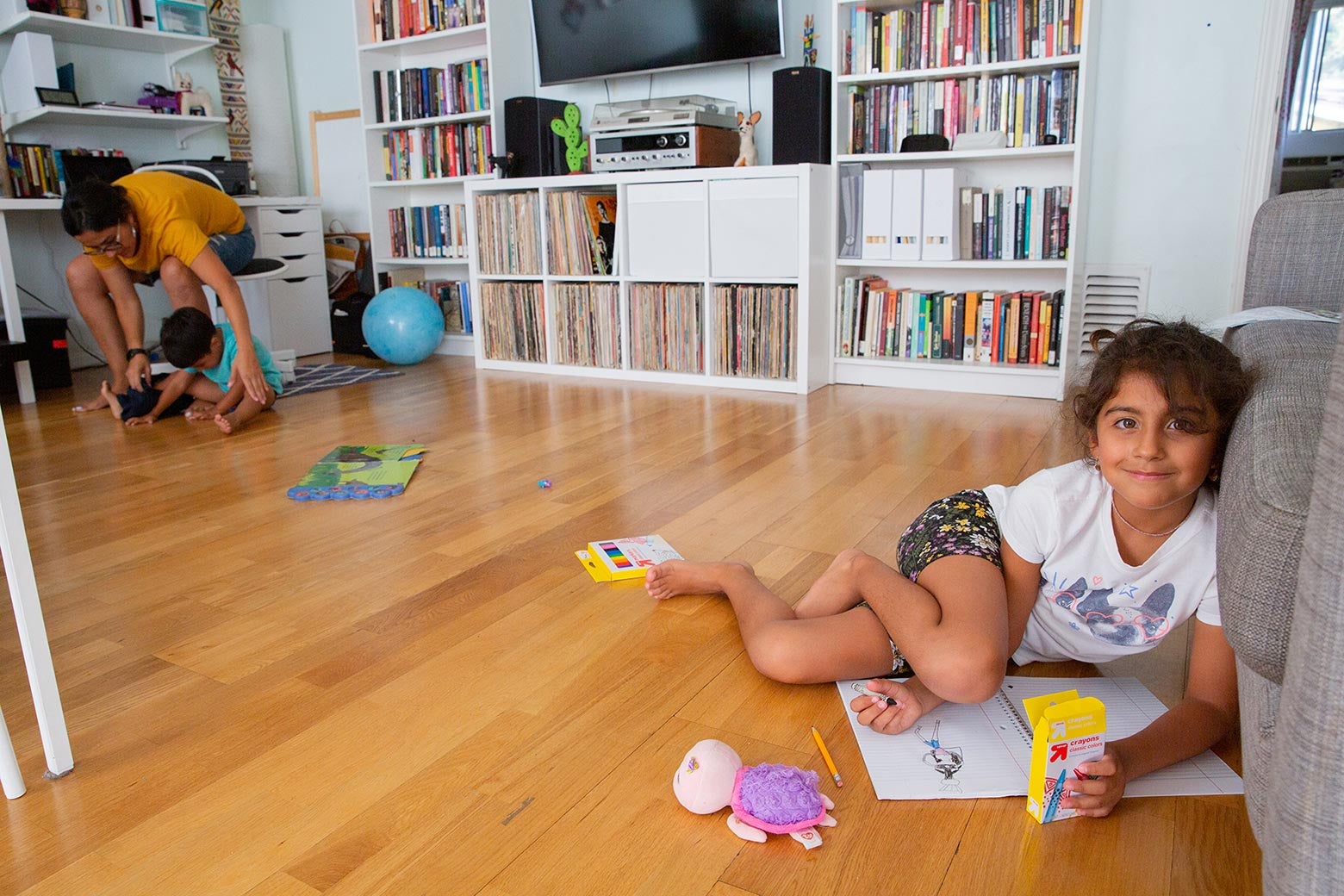 A little girl sitting on the ground with her art supplies smiles at the camera. On the other side of the room, her mother reaches down to her son, who's sitting on the floor. In the background are bookshelves and a TV.