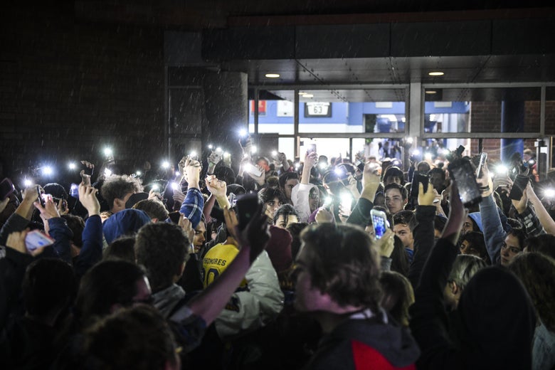 Students hold up their lit phones as they exit a high school.