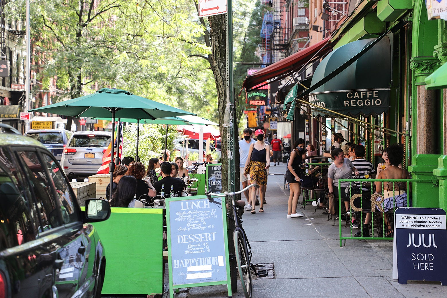A busy street: sidewalk cafe diners, menu boards, a sign for Juul, and pedestrians.