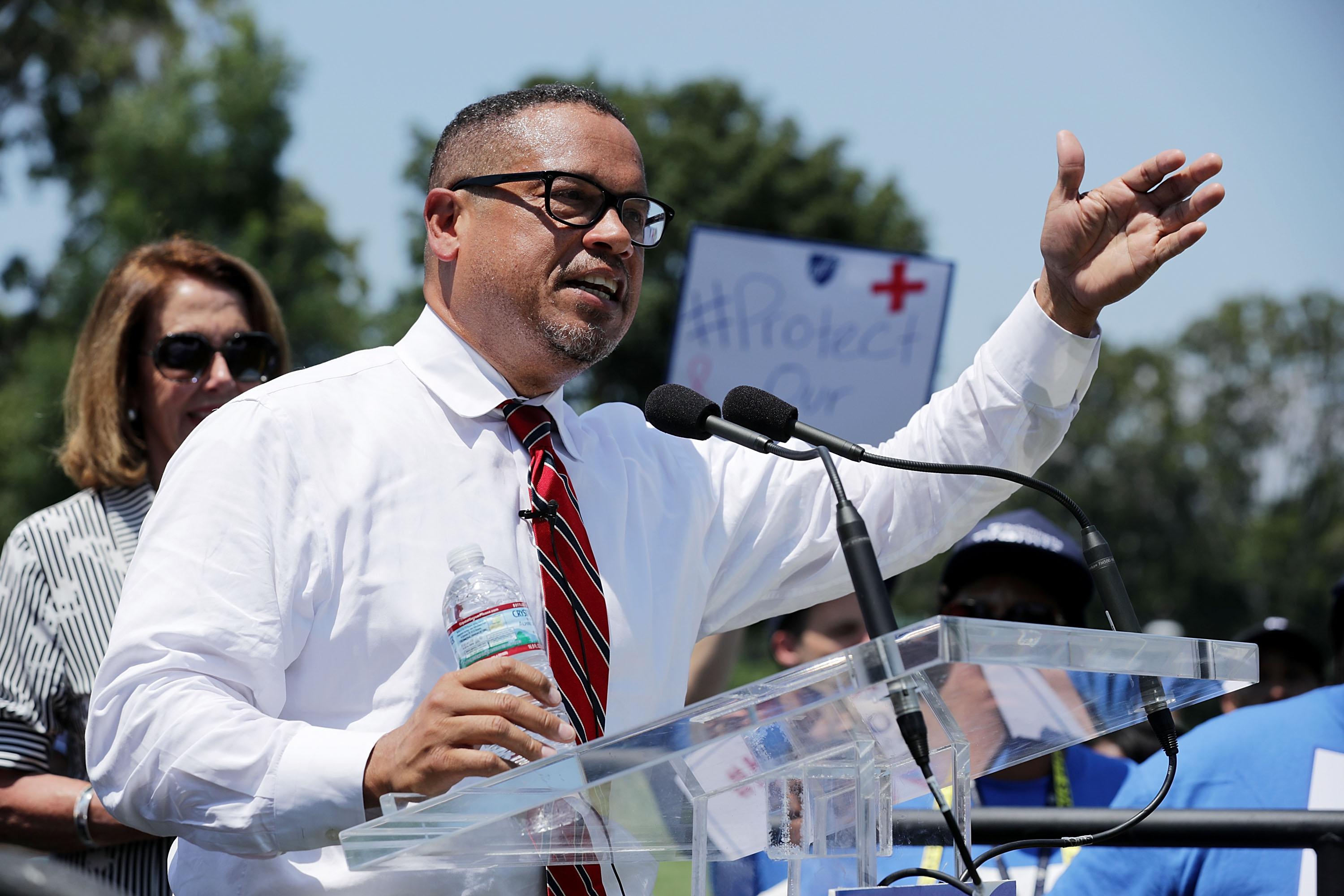 Ellison, in a white shirt and red tie, raises his arm to gesture to a crowd as he speaks. Behind him, people wave signs.