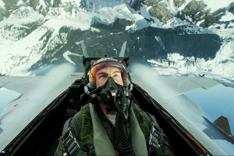 Tom Cruise wearing a flight helmet in the cockpit of a fighter jet that is upside-down over a mountain in a scene from Top Gun: Maverick