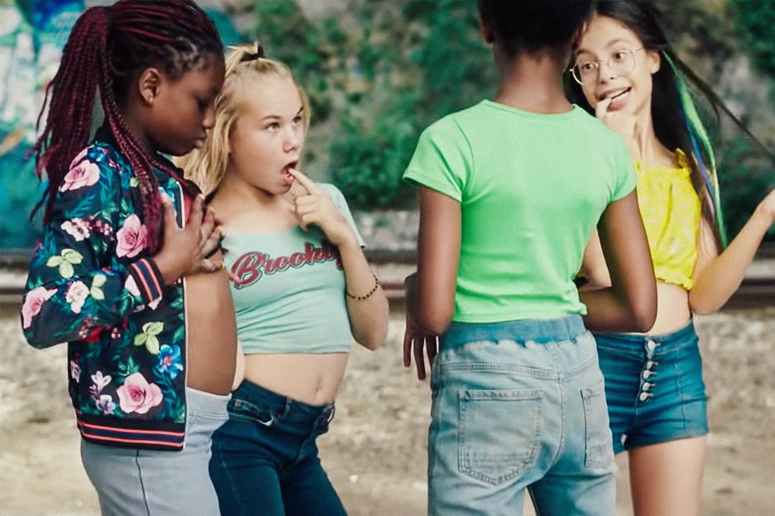 Four tween girls stand around, as one puts her finger in her mouth and another puts her hands on her chest.
