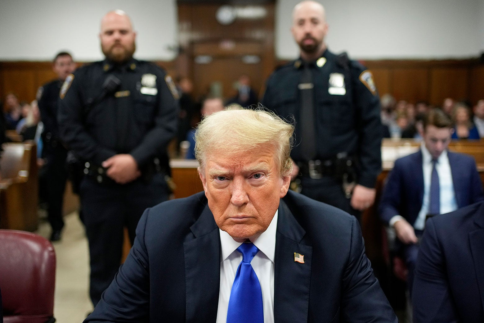 Trump glares at the camera, two officers stand behind him and in front of the rest of the courtroom.