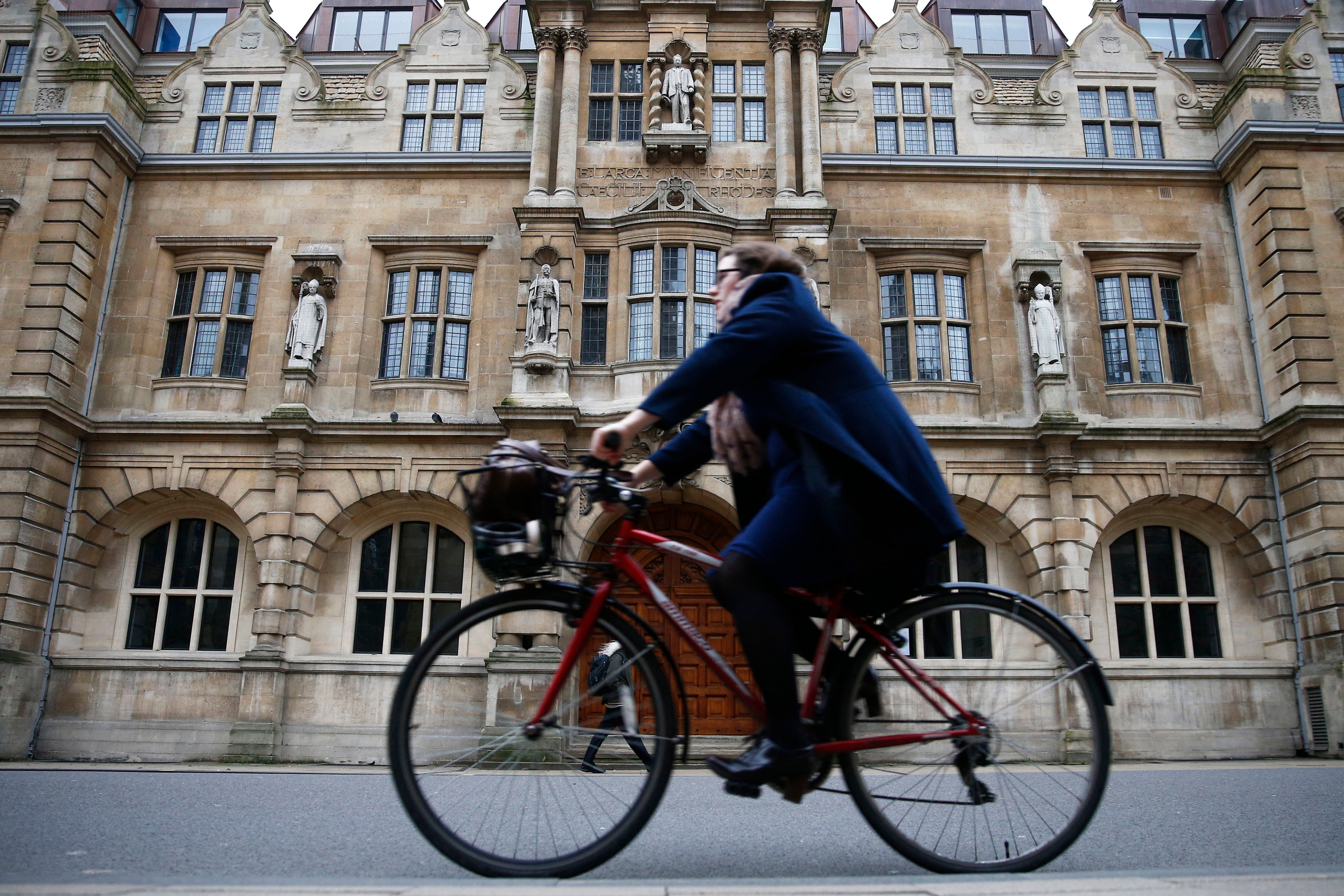 A woman bicycles in front of an old building in Oxford, England. She is wearing a blue coat.