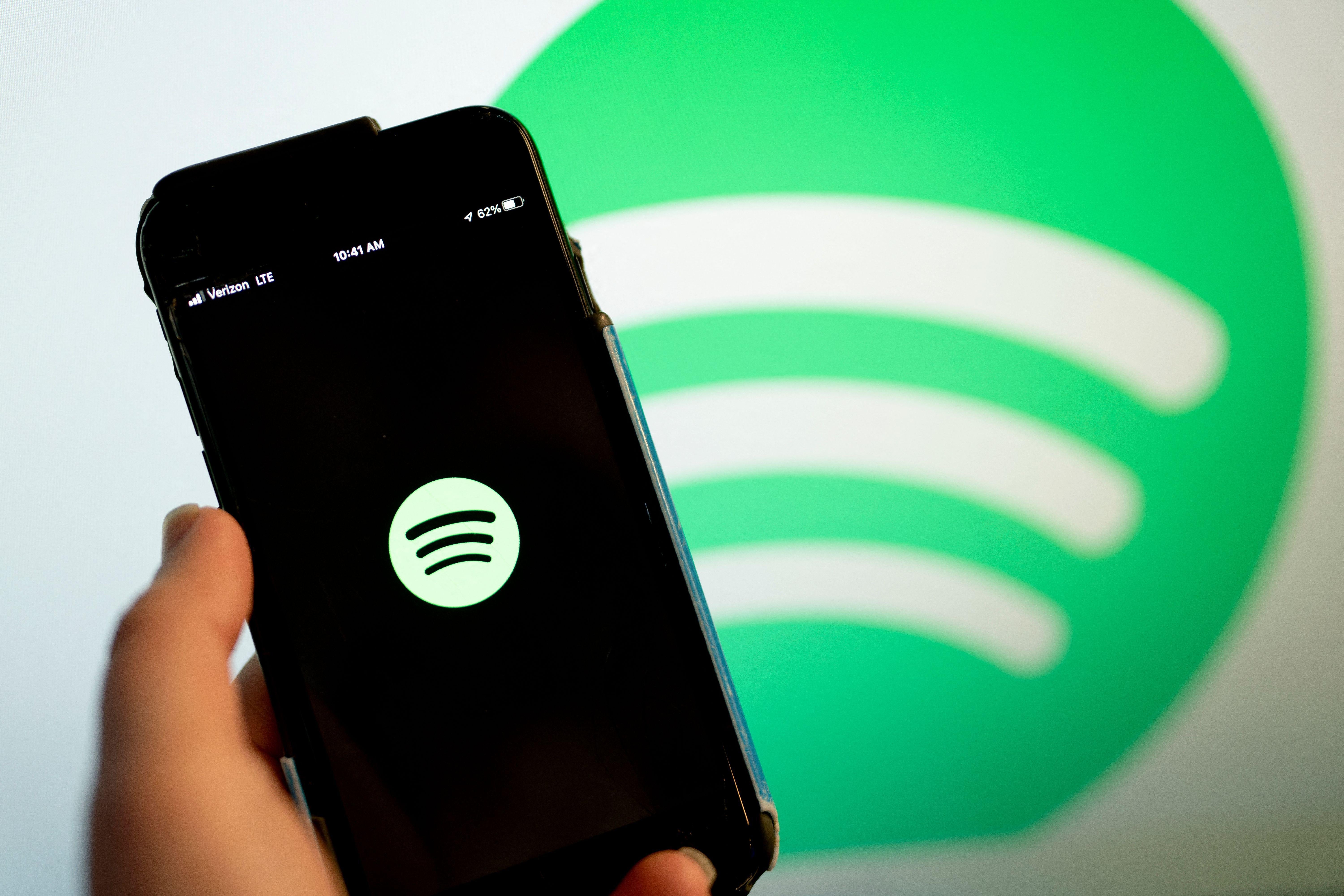 In front of a large Spotify logo, a hand holds a smartphone that displays another Spotify logo.