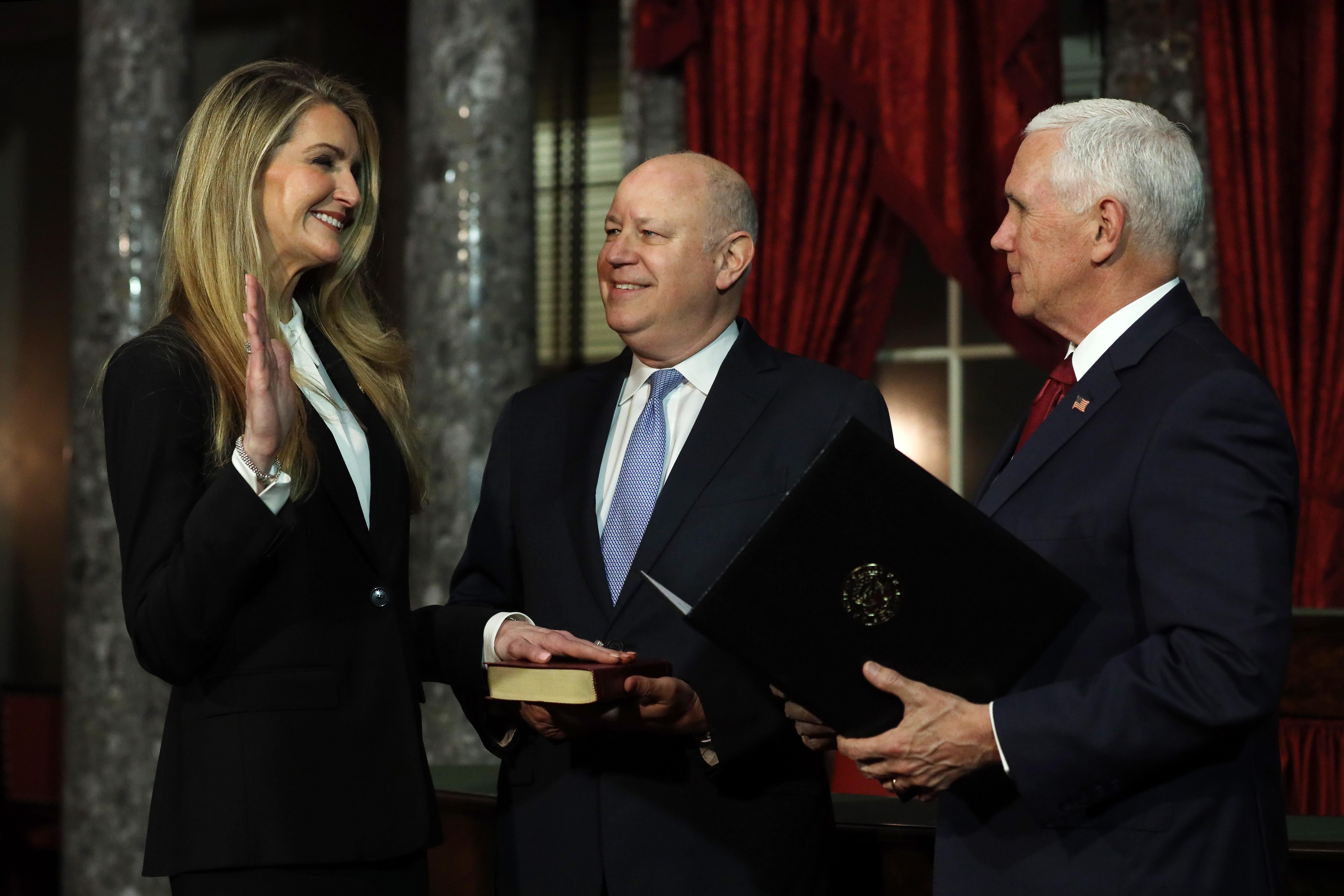 Loeffler smiles and holds up her right hand as she puts her left hand on a Bible held by her husband, with Pence standing beside them.