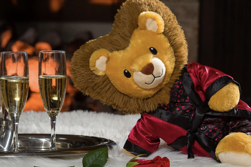 A smiling stuffed lion in a red silk robe reclines on a white carpet beside a glass of champagne