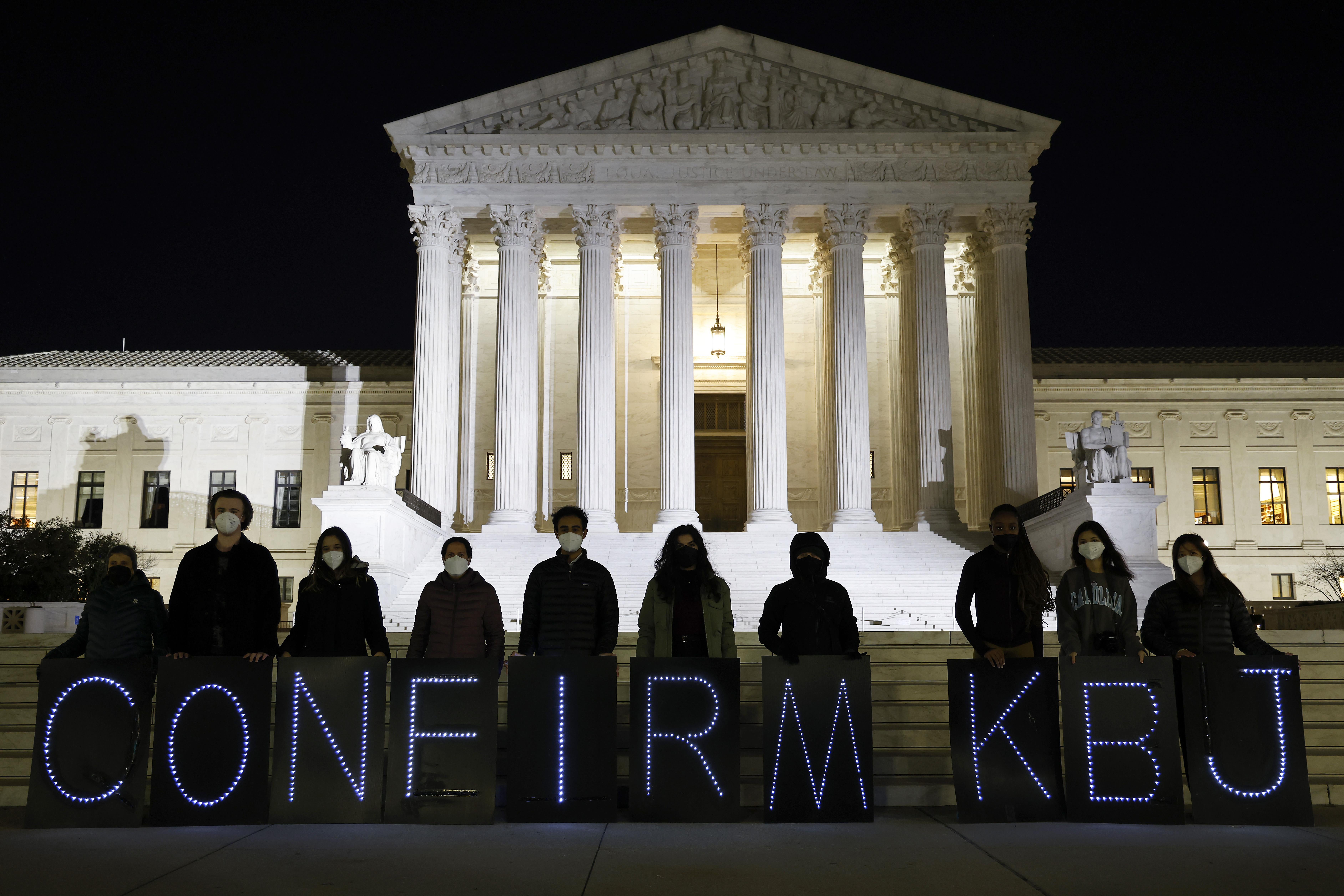 People stand in front of the Supreme Court at night holding up signs with letters spelling out "CONFIRM KBJ"