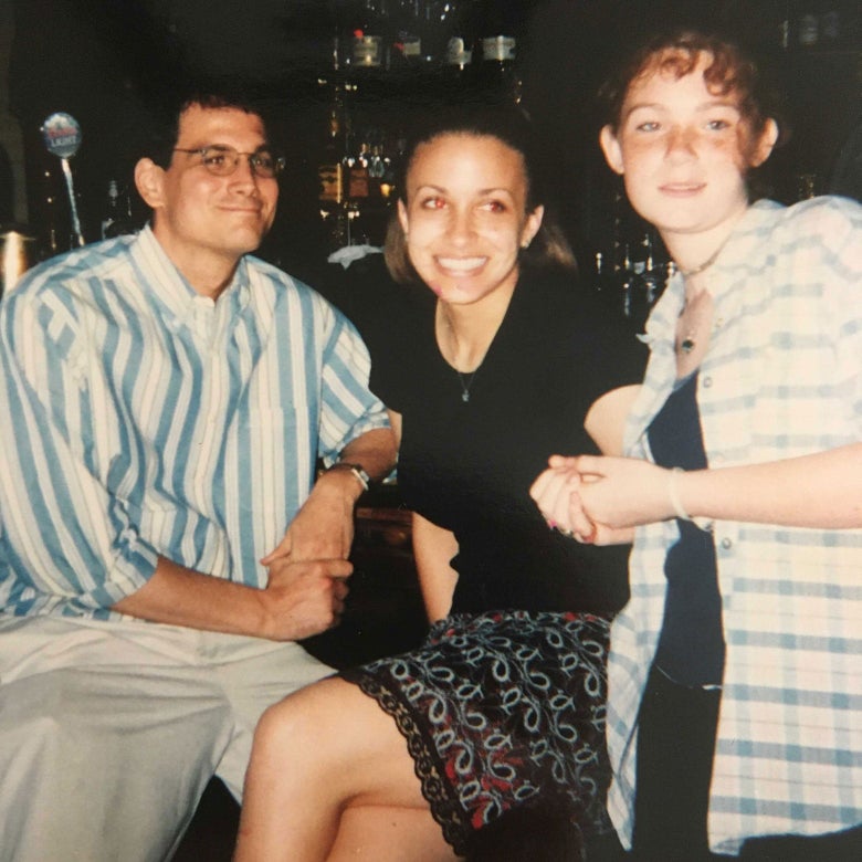 A man in a stripped button down and khakis smiles as he sits on a bar stool next to two teenage girls.