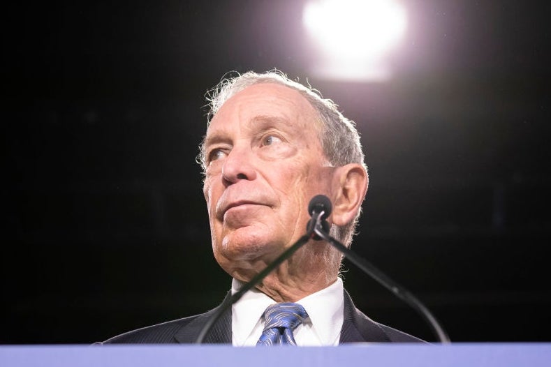Bloomberg, standing at a blue lectern, looks to his right against a black backdrop.