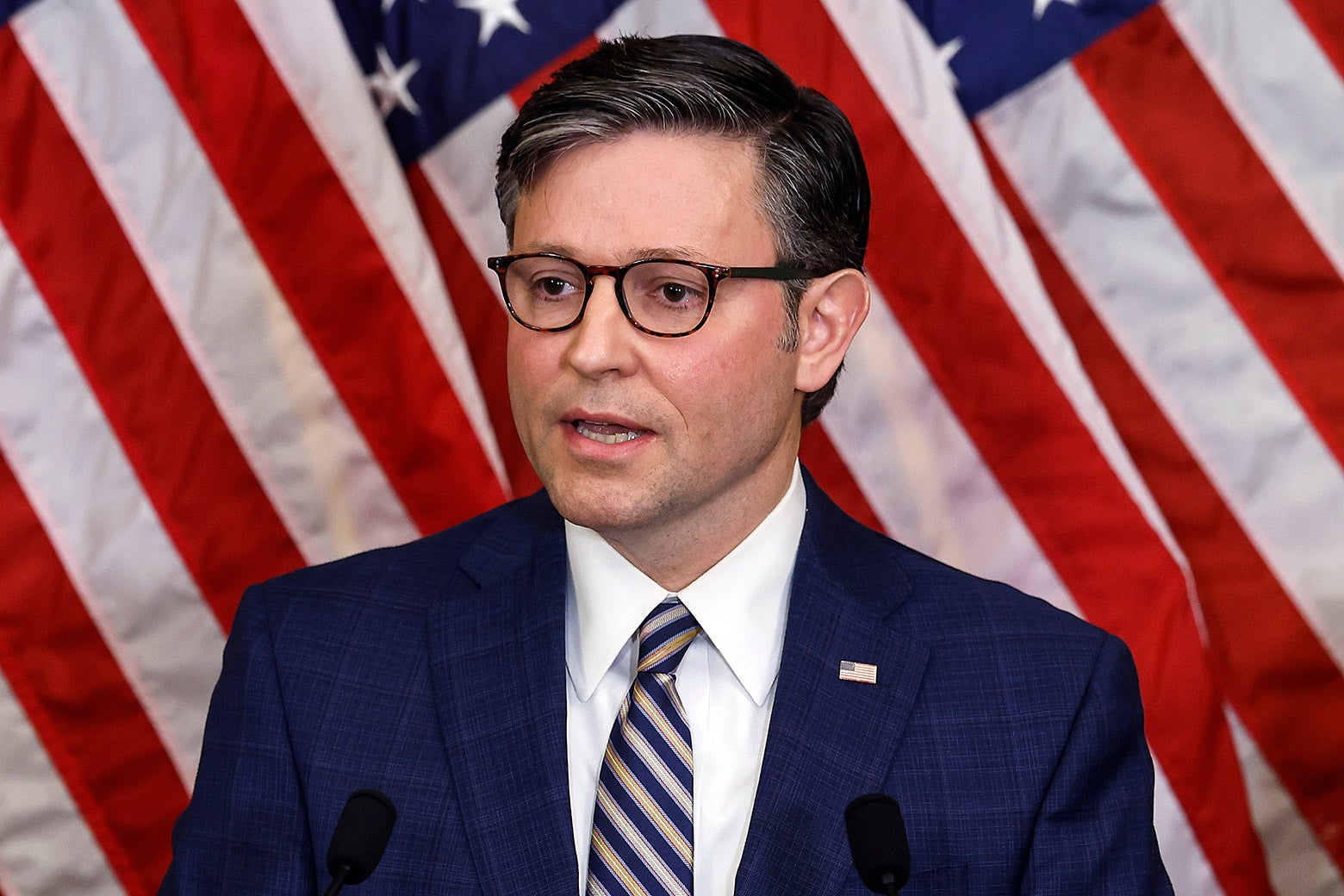 Speaker of the House Mike Johnson appears in front of an American flag. He is wearing Clark Kent–style glasses, and his mouth is slightly agape.