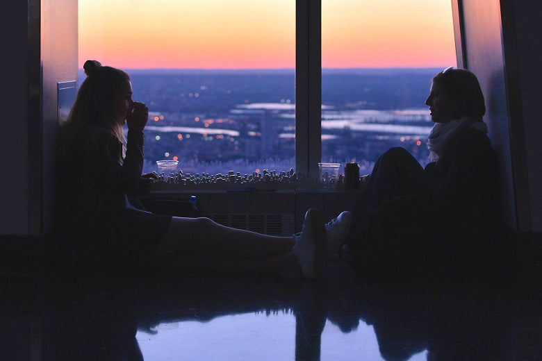 Two women having a discussion while sitting on the floor against the backdrop of a sunset through a large window.