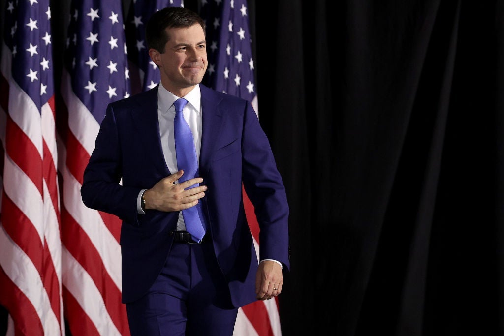Buttigieg, smiling and wearing a blue suit, strides in front of American flags onstage.