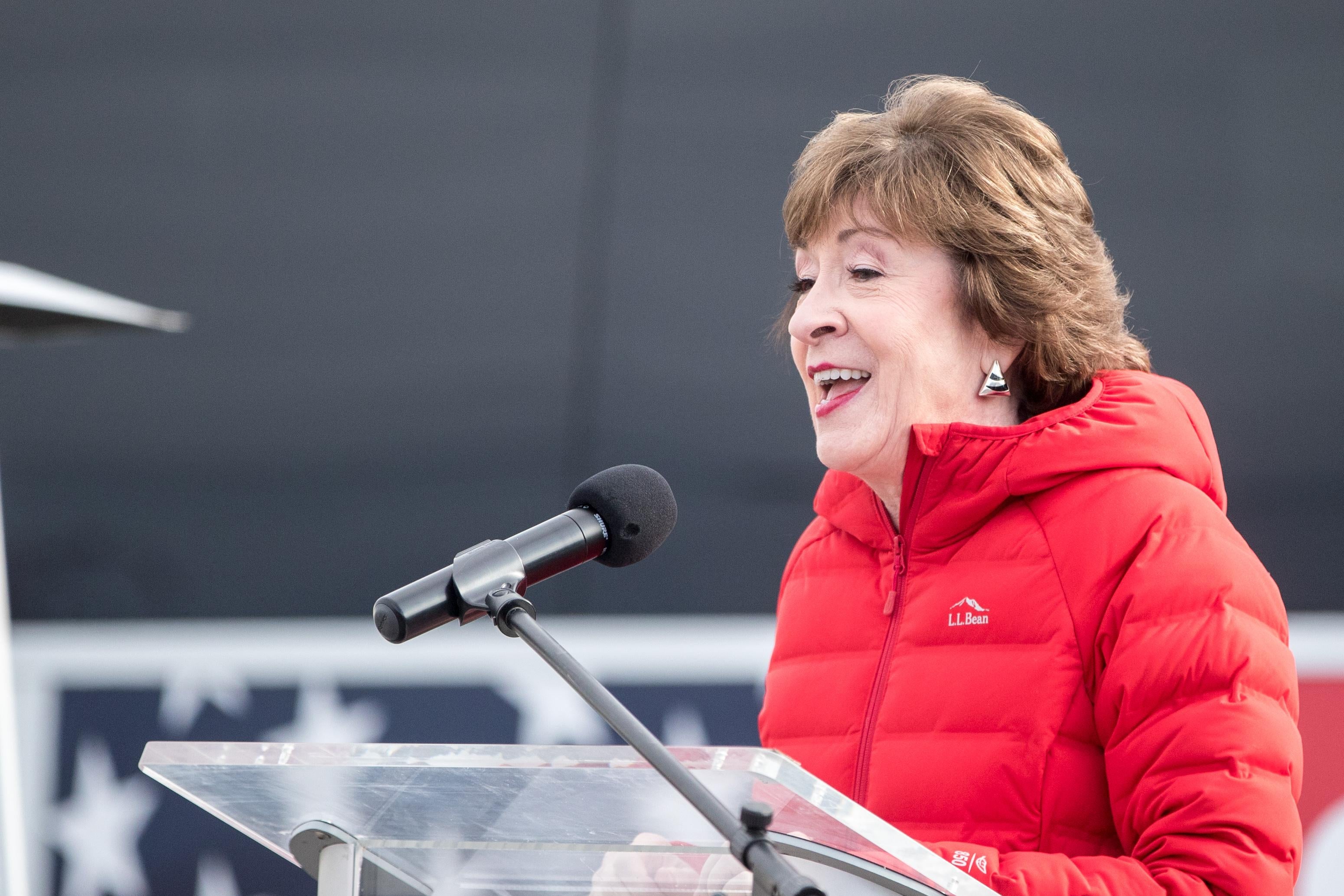 Susan Collins, in a red parka, speaks into a microphone at a transparent podium.