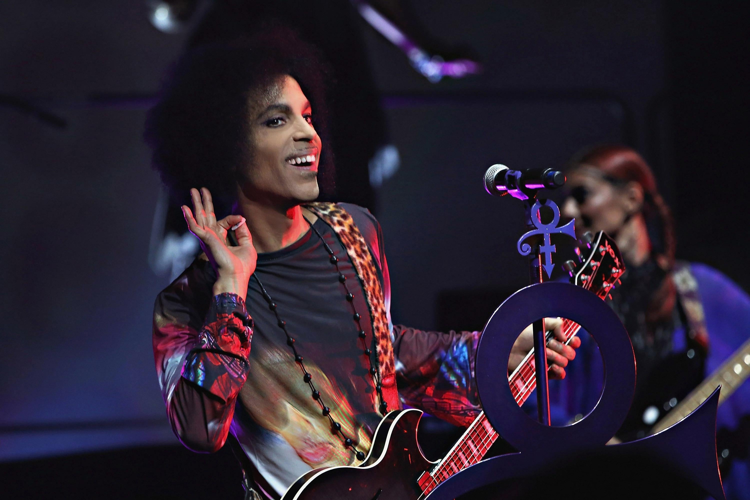 A man with a large afro and wearing a multi-colored suit holds a guitar on a stage. He leans to the right side of the microphone with his hand by his ear, smiling. 