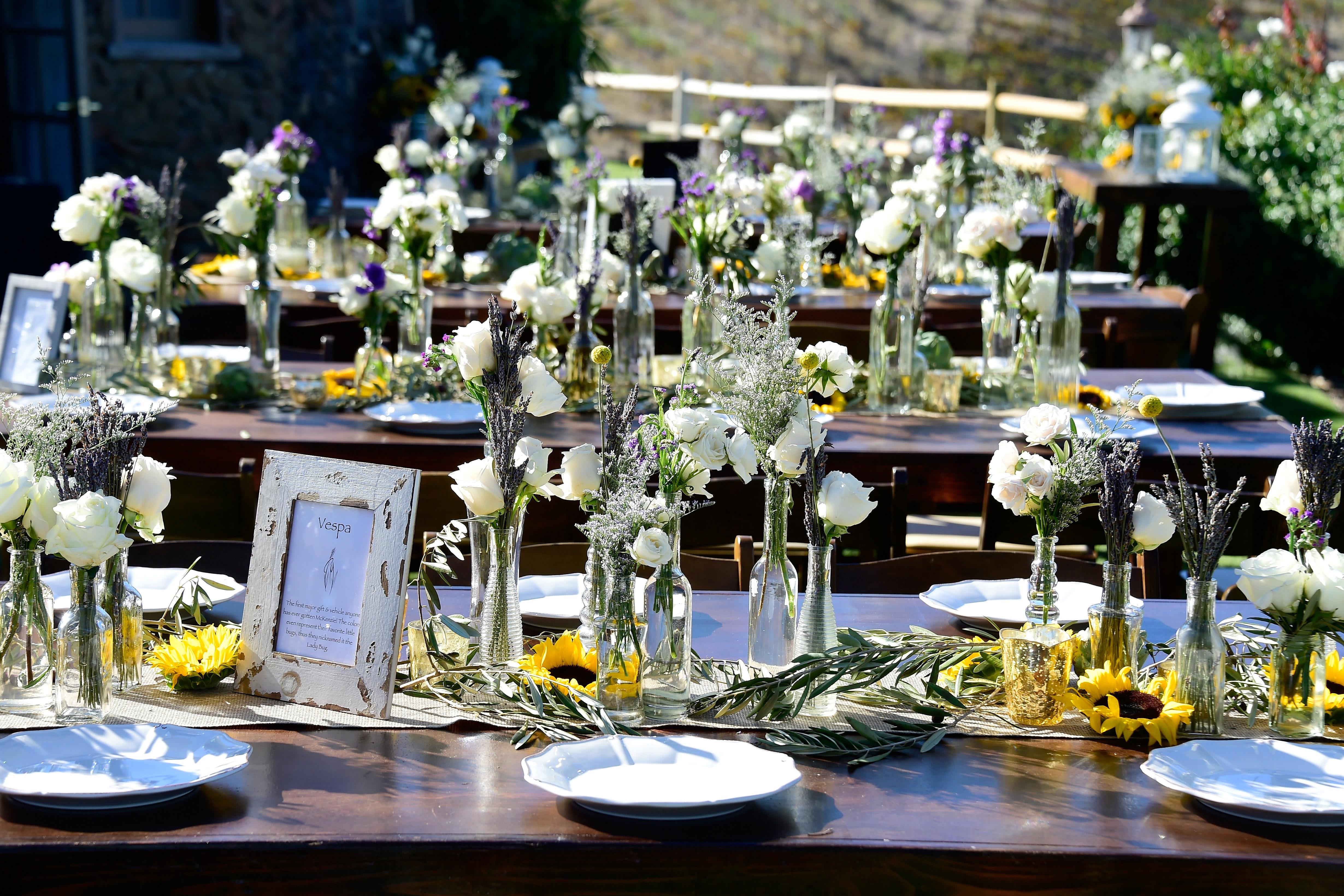Flowers in vases and porcelain plates sit on dark wooden benches at an outdoor wedding 