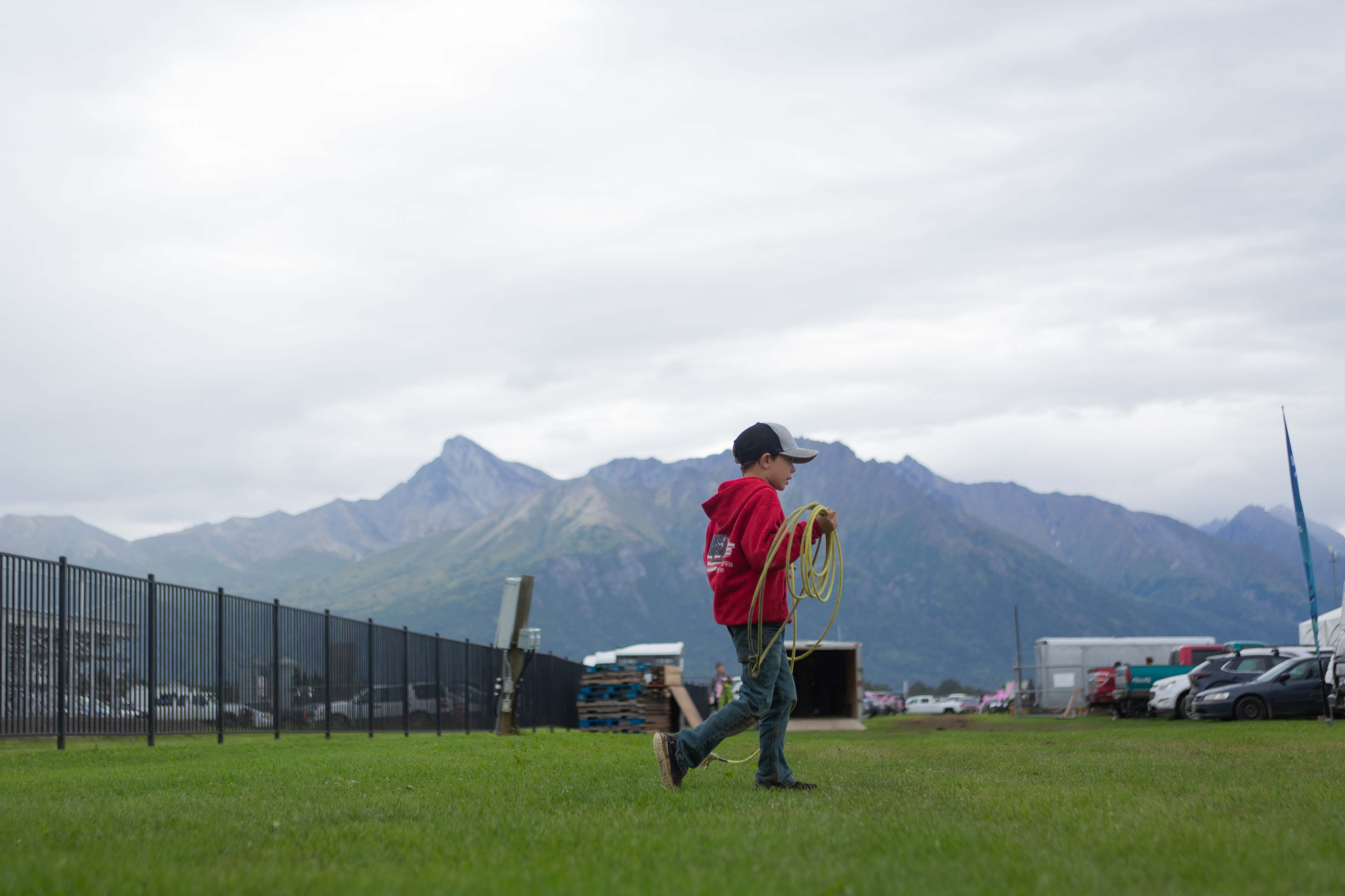 A boy holding a lasso walking across a field with the Alaskan mountains in the background.
