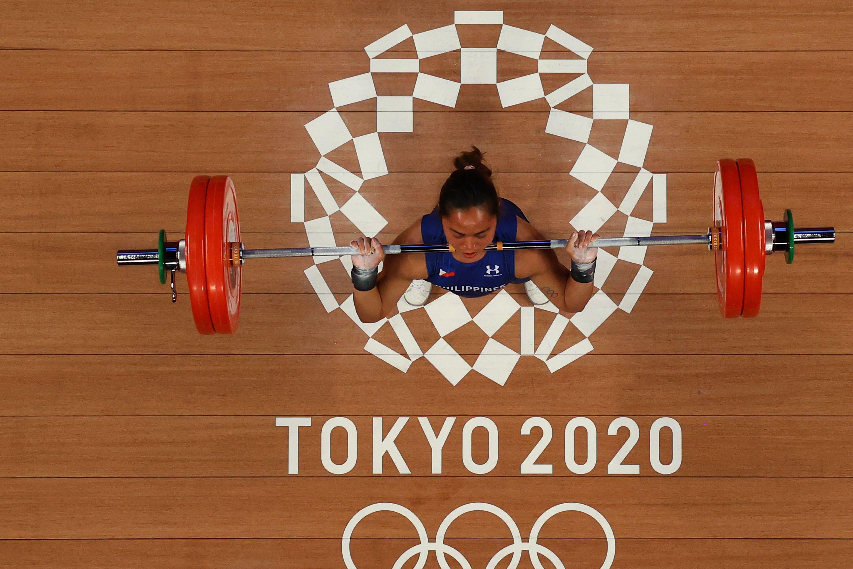 An overview of a female weightlifter midway through a barbell lift, with the Tokyo 2020 Olympics logo underneath her feet.