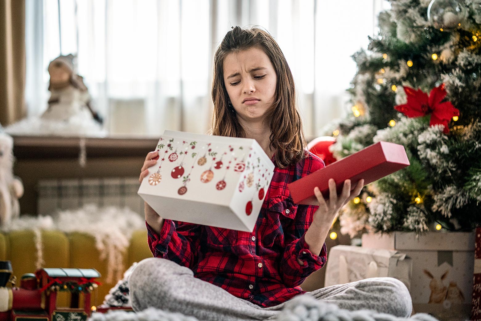 A girl looks skeptically into a Christmas box near a Christmas tree.