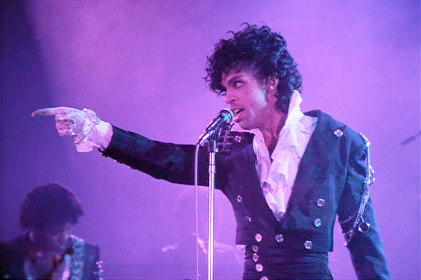 The musician Prince stands on stage in a white ruffled shirt, backlit by purple stage lights, and points to someone in the crowd. 