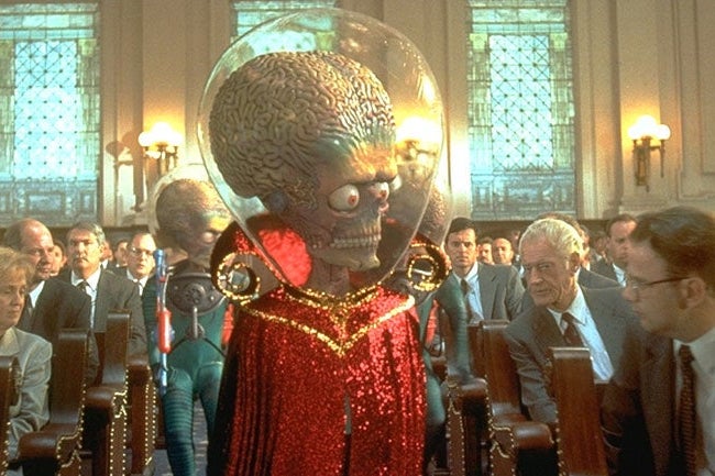 An alien with a giant head, wearing a glittery red robe and bubble helmet, walks down an aisle surrounded by people in suits.