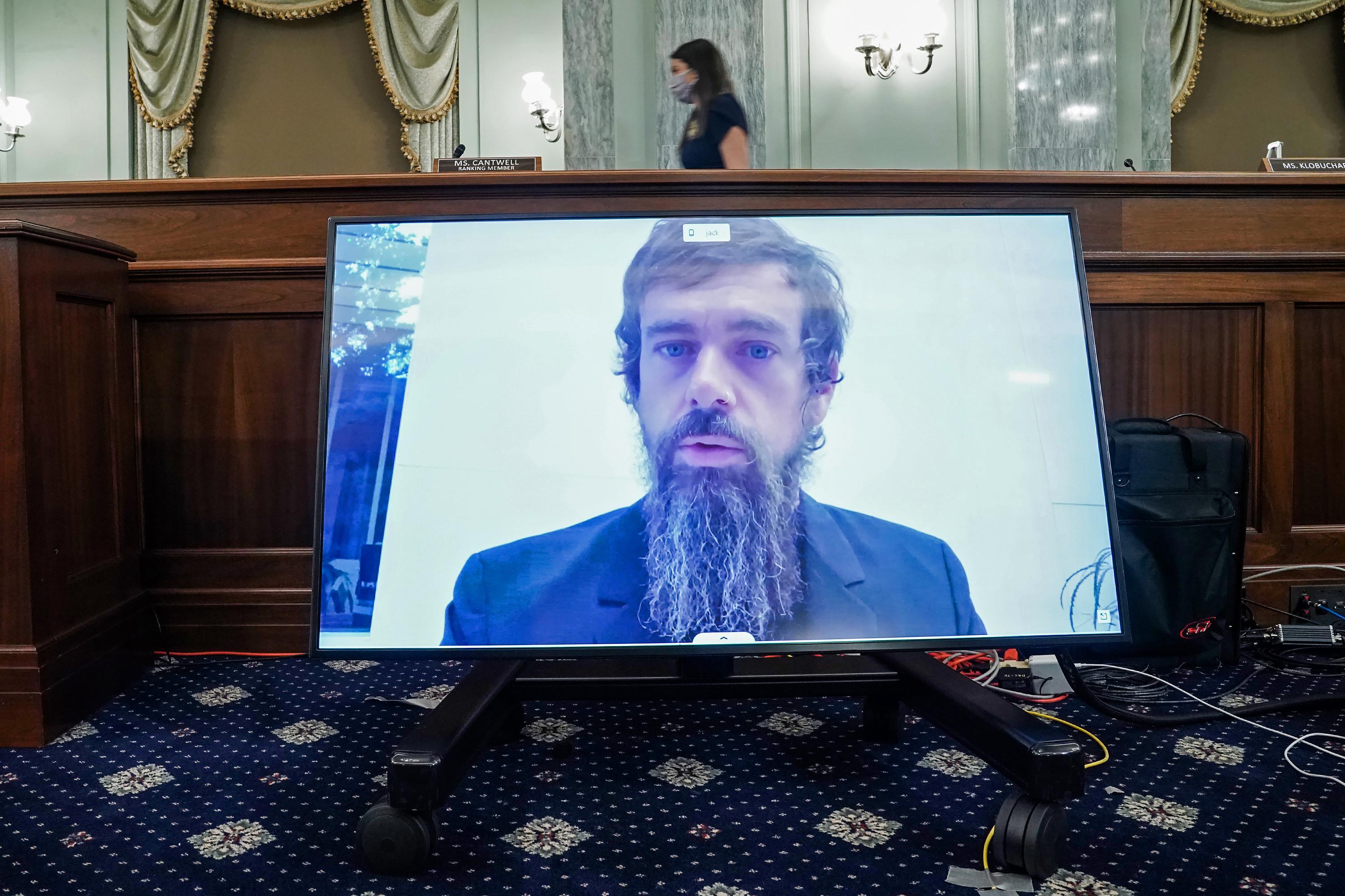Jack Dorsey appears with unkempt hair and a long, scraggly brown and gray beard on a screen sitting on the floor of a Senate hearing room