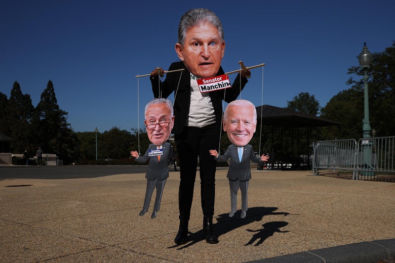A demonstrator wears a photograph of Sen. Joe Manchin as a mask while playing a puppet-master, holding a puppet of Chuck Schumer in one hand and Joe Biden in the other. 
