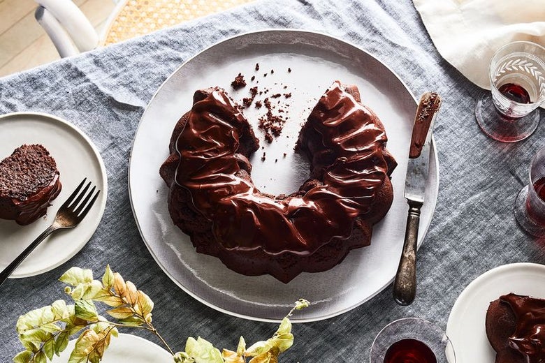A decadent chocolate cake, covered in ganache, sits at the center of a table. A piece is cut out and sits on a plate to its left. There is a glass of Passover wine on its right.