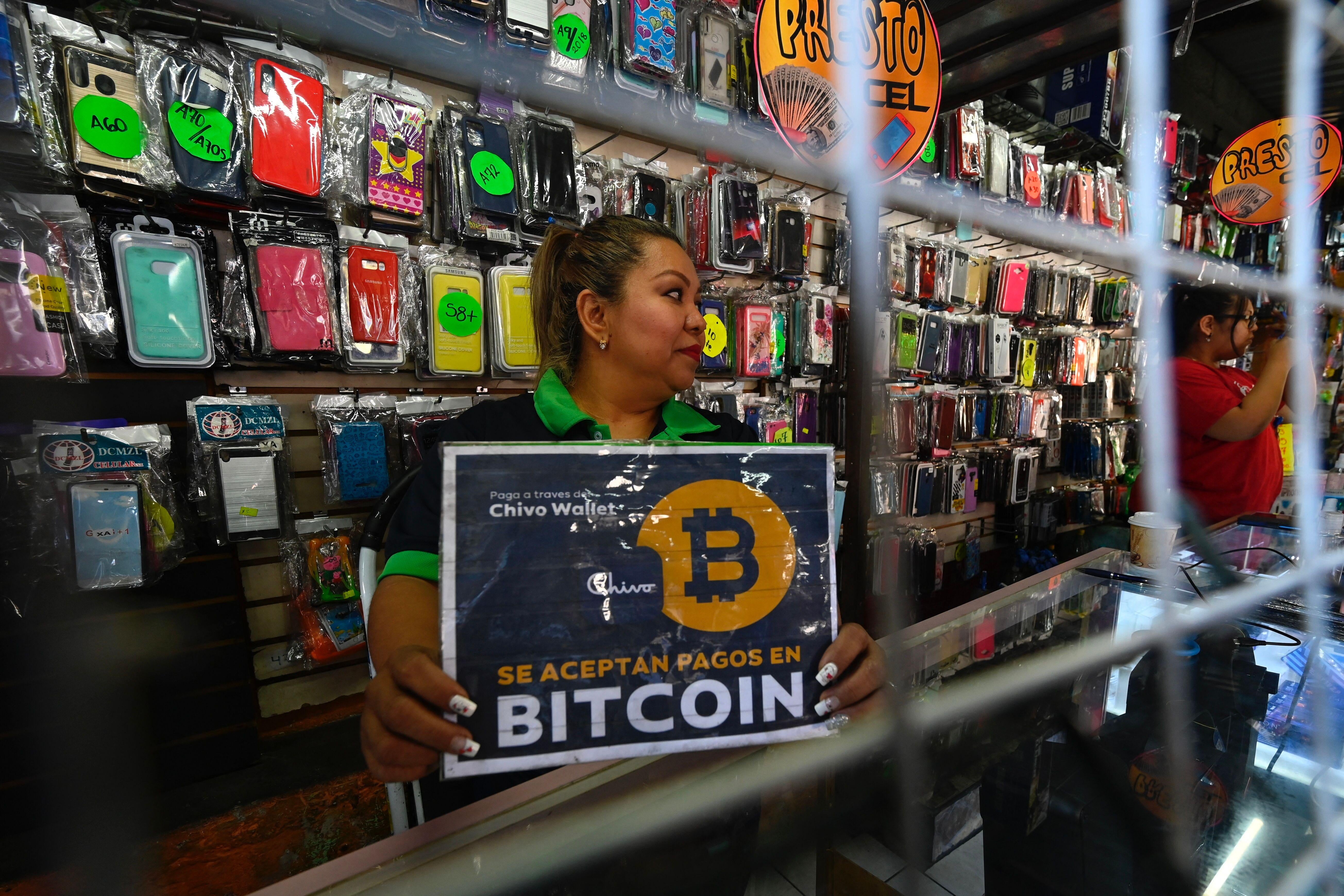 A woman holds a sign saying "Bitcoin Accepted" at a counter in front of a wall of products.