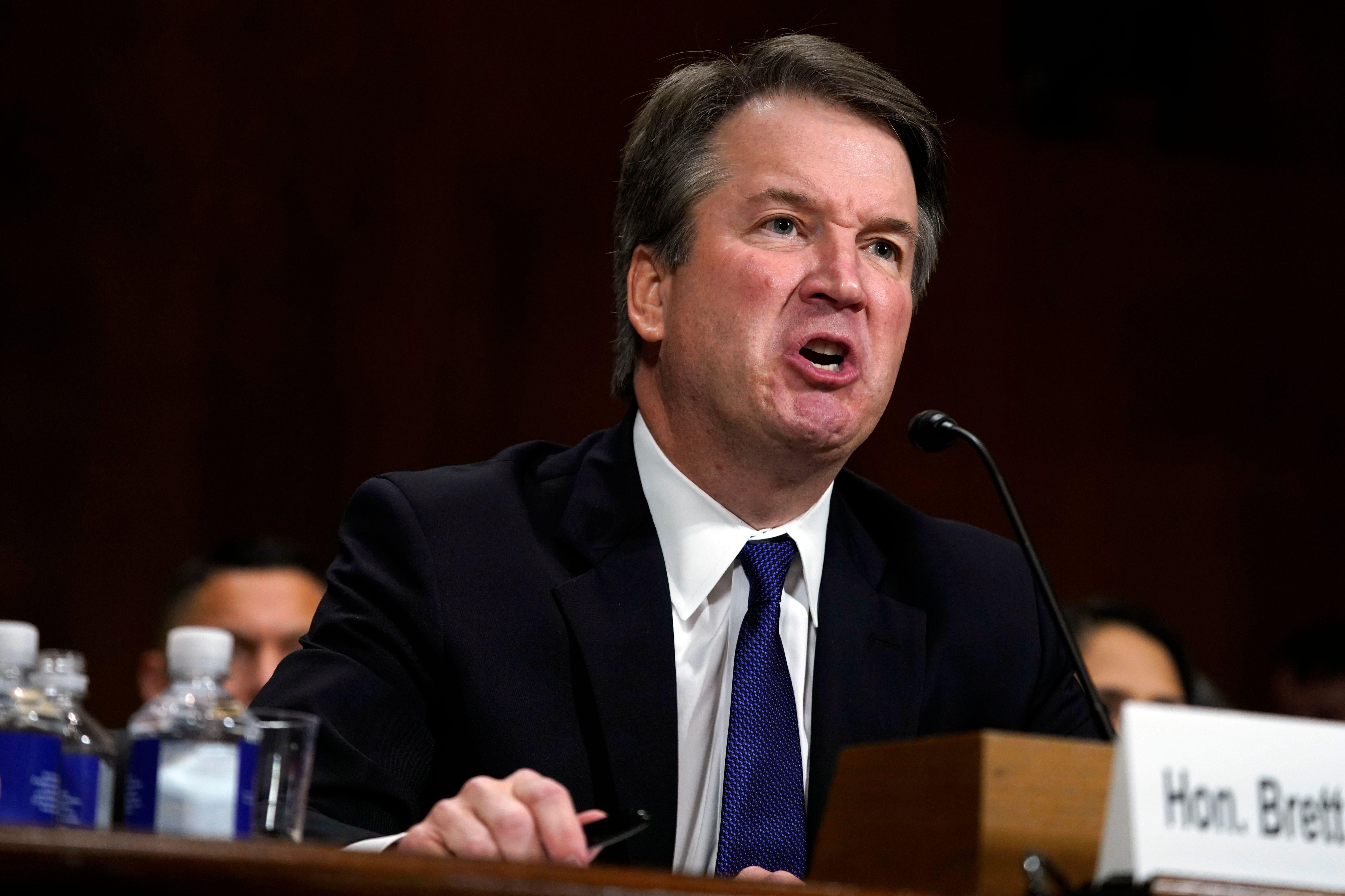 Brett Kavanaugh scrunches up his face as he shouts into the microphone on a table. 