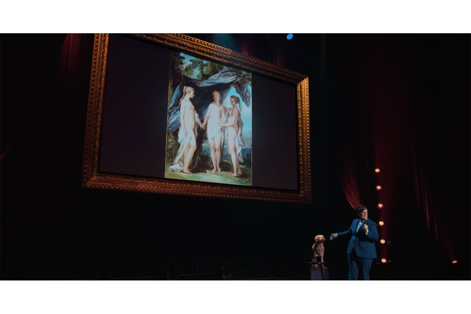 Hannah Gadsby onstage in front of a painting of three naked women.