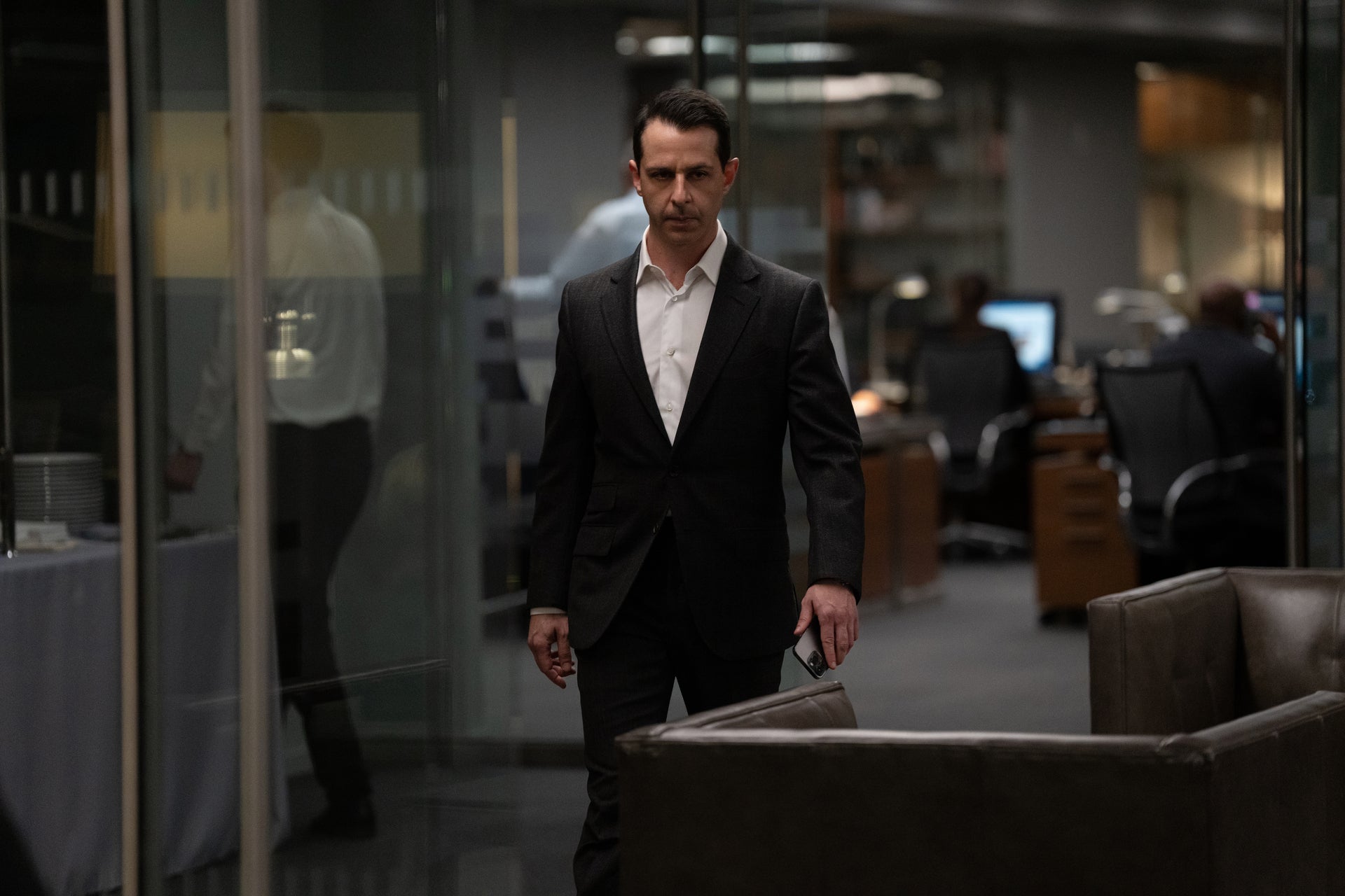 In a black suit and an unbuttoned white shirt, he struts confidently but somberly down an office hallway.